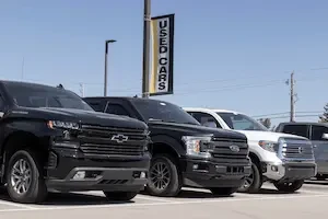 A row of used cars in a lot under a used cars sign. There are two large black SUVs or trucks, a white one, and a silver one.