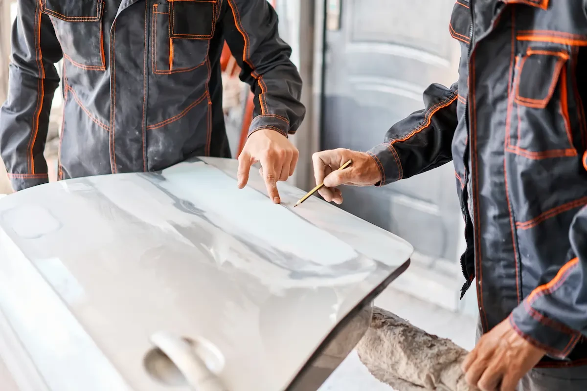 Two auto body technicians in dark work uniforms with orange stitching examine a white vehicle body panel on a workstation. One points at a section of the panel while the other holds a pencil marker near the surface.