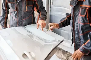 Two auto body technicians in dark work uniforms with orange stitching examine a white vehicle body panel on a workstation. One points at a section of the panel while the other holds a pencil marker near the surface.