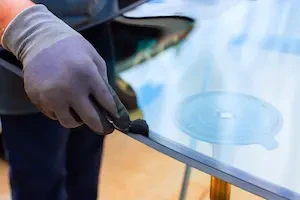 A gloved technician's hand applies primer to the edge of a windshield with a small foam applicator, preparing the glass for installation on a stand in an auto glass shop.