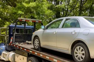 A silver sedan is secured with wheel chocks on the deck of a dark blue flatbed tow truck, parked on a tree-lined road