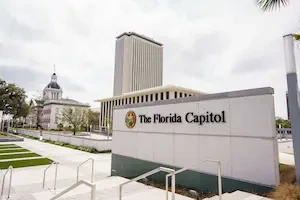 A sign reading "The Florida Capitol" stands in front of a plaza, with the modern high-rise capitol tower and the historic domed capitol building visible in the background.