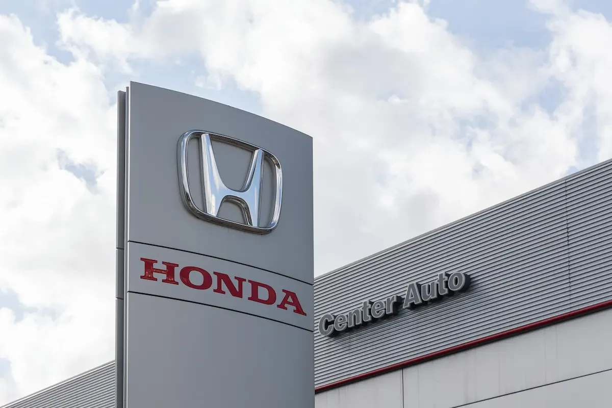 A Honda dealership pylon sign with the Honda logo and "HONDA" in red lettering against a cloudy sky, with a "Center Auto" building sign visible in the background.