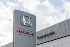 A Honda dealership pylon sign with the Honda logo and "HONDA" in red lettering against a cloudy sky, with a "Center Auto" building sign visible in the background.