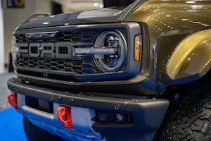 Close-up of an olive green Ford Bronco Raptor's front grille and headlight, with an orange tow hook on the bumper, photographed indoors on a blue floor covering at an auto show.