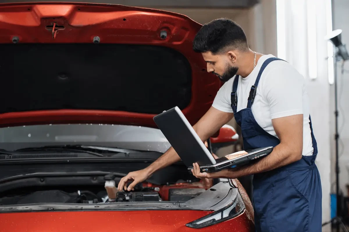 collision repair tech standing by open hood hood of red car with a laptop