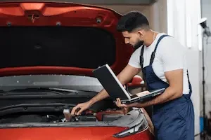 collision repair tech standing by open hood hood of red car with a laptop