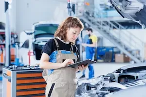 Technician in tan coveralls uses a tablet to calculate an invoice in a repair shop, with a rolling tool cart beside her and a car with its hood open in the foreground; a second technician in blue coveralls reviews a clipboard in the background.