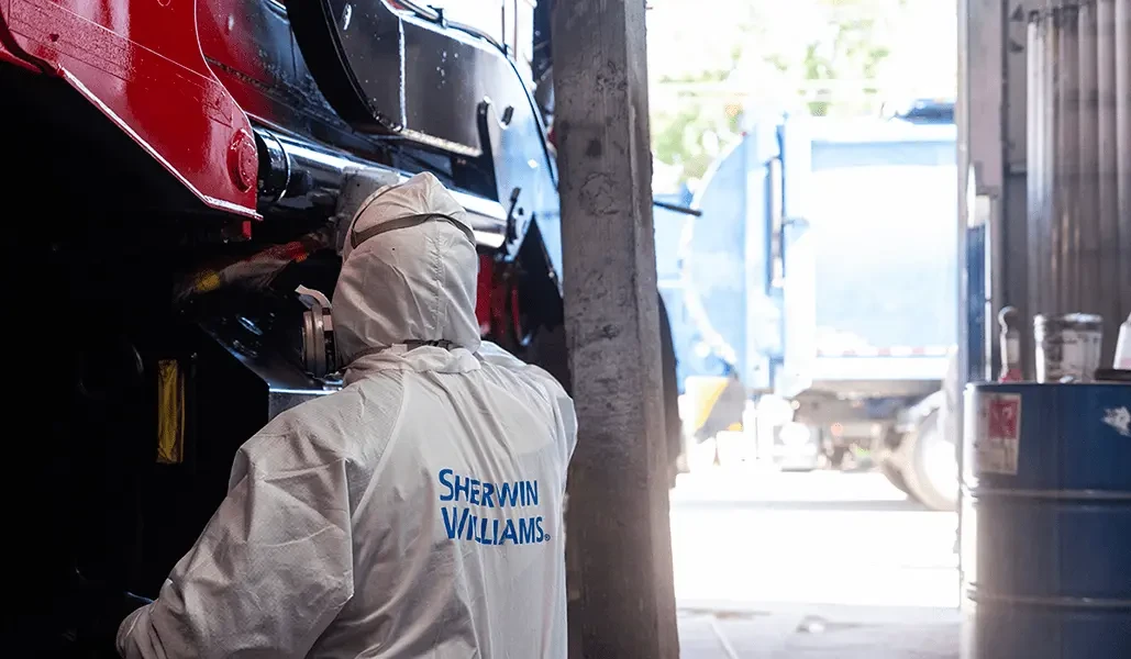 Technician wearing a white protective coverall with a Sherwin-Williams logo and respirator mask works underneath the raised front end of a red commercial truck in a shop bay, with a garbage truck visible outside.