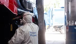 Technician wearing a white protective coverall with a Sherwin-Williams logo and respirator mask works underneath the raised front end of a red commercial truck in a shop bay, with a garbage truck visible outside.