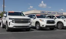 A row of white SUVs, including a Chevrolet and a Toyota, parked in a used car dealership lot with price stickers on the windshields showing figures such as 72989, 20534, and 28689. A sign reading "USED CARS AND TRUCKS" is visible on the building in the background under a partly cloudy sky.