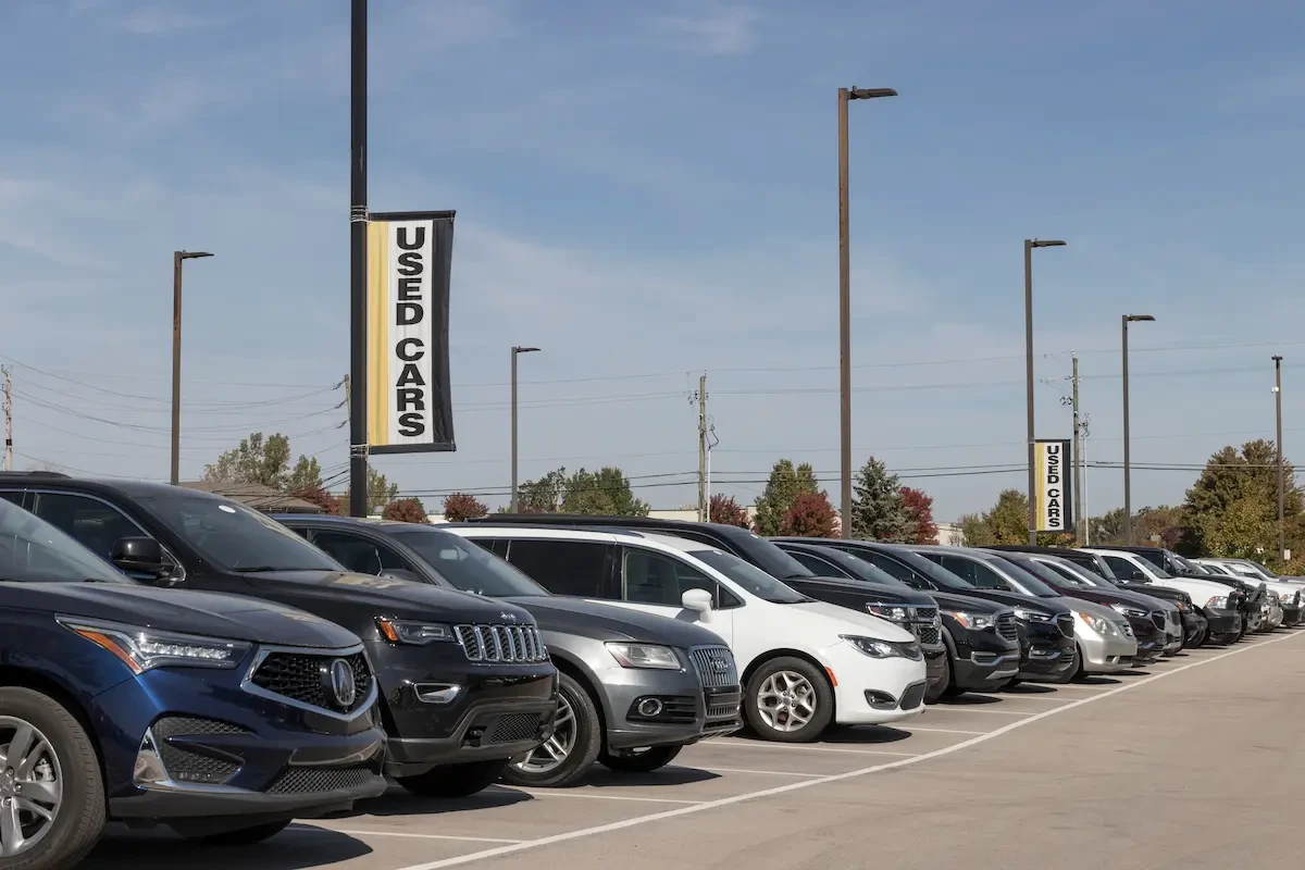 Rows of used SUVs and sedans, including an Acura, Jeep, Audi and Chevrolet, parked at a dealership lot beneath tall light poles, with a black, white and yellow "Used Cars" banner hanging above and trees and power lines visible in the background under a partly cloudy sky.