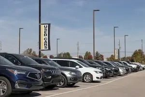 Rows of used SUVs and sedans, including an Acura, Jeep, Audi and Chevrolet, parked at a dealership lot beneath tall light poles, with a black, white and yellow "Used Cars" banner hanging above and trees and power lines visible in the background under a partly cloudy sky.
