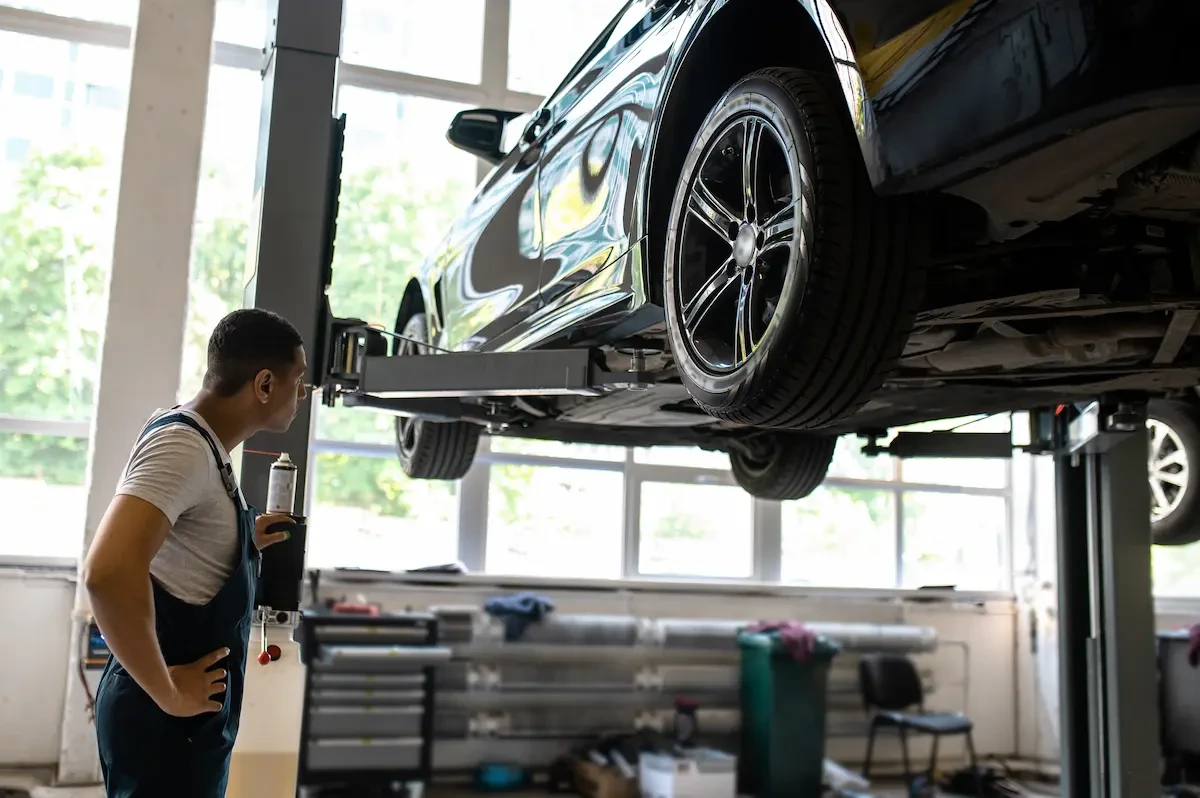 A technician in coveralls stands beside a vehicle raised on a two-post lift, holding a spray canister and looking up at the vehicle's underside in an auto repair shop.