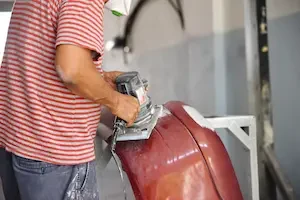 A technician in a red-and-gray striped shirt uses a handheld orbital sander on a maroon vehicle panel, with white sanded patches visible where the finish has been feathered down. The panel is removed from the vehicle and mounted on a metal stand.