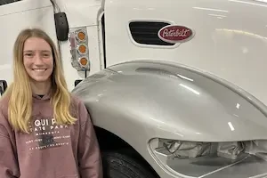  A young woman smiling in a collision repair shop, standing next to a painted silver truck fender.