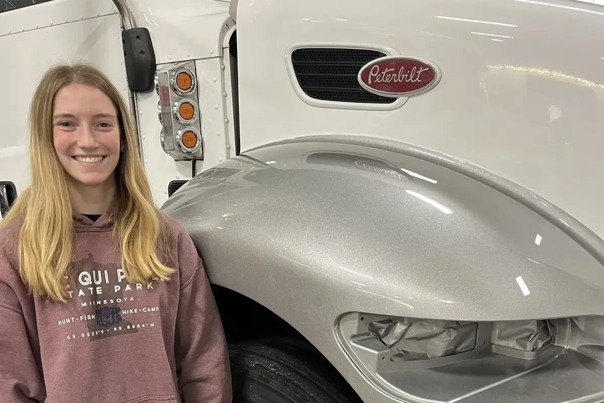 A young woman smiling in a collision repair shop, standing next to a painted silver truck fender.