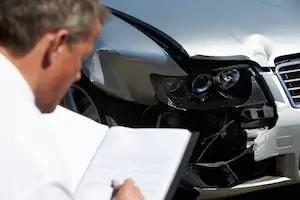 man writing in a notebook in front of a damaged front of a car