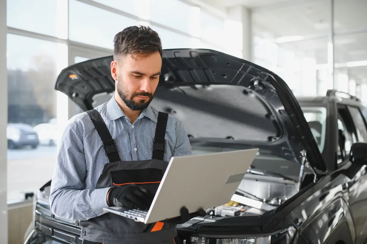 A man with a laptop standing in front of a car with an open hood 