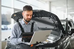 A man with a laptop standing in front of a car with an open hood 