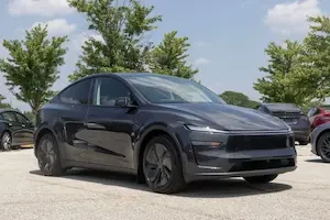 Dark charcoal Tesla Y car in a parking lot with green trees and a blue sky