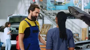 A white man with dark hair and a beard talking to a black woman with dark hair in braids wearing a blue with her back to the camera. They are in front of a car with its hood raised with another woman in front of a car with the trunk open in the background.