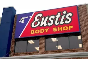 Exterior of a red brick building with a blue chimney, two large windows, and a red and blue sign that says Eustis in white and body shop in yellow