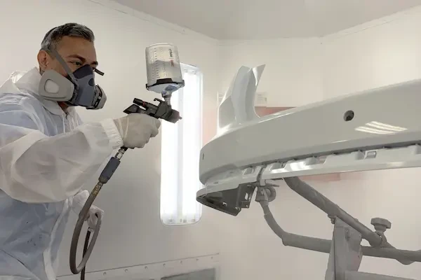 Man in white PPE and respirator spraying a car bumper in a paint booth