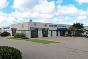 Exterior of white rectangular building with blue accents and two garage bays with the sign Zimbrick Body Shop on the front in blue.
