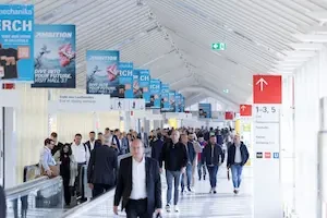 People walking in a brightly lit conference center hallway with informational/directional red and white signs to the right of the walkway and blue promotional banners hanging down from the ceiling.
