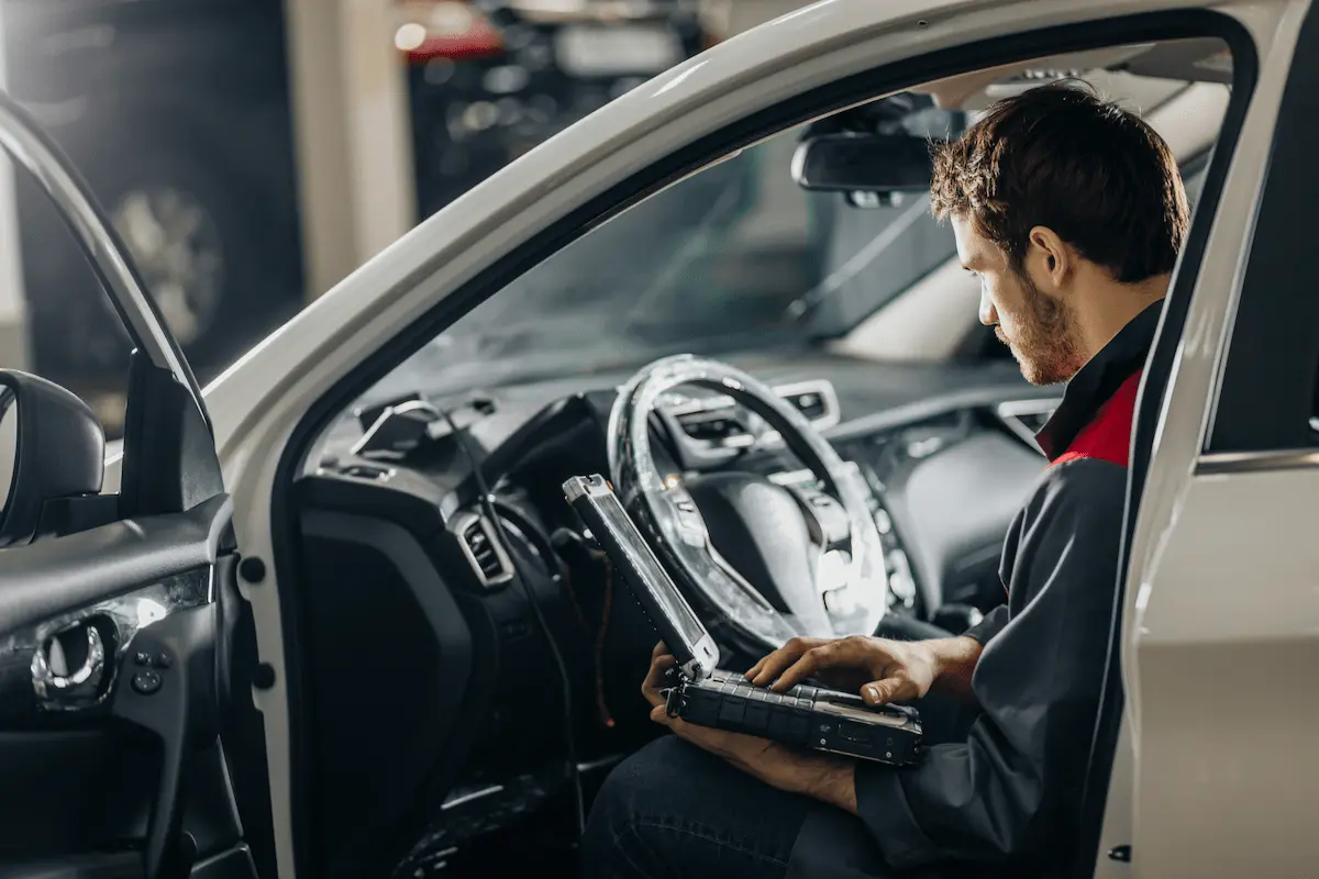 Technician seated in a vehicle’s driver’s seat using a rugged laptop connected to the car for diagnostics.