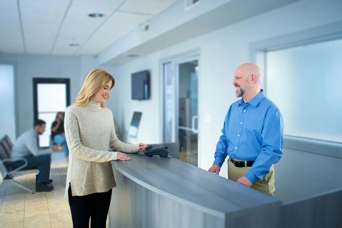 A collision repair shop customer pays for a repair at the front desk while a service advisor assists, illustrating the cost and payment conversation.