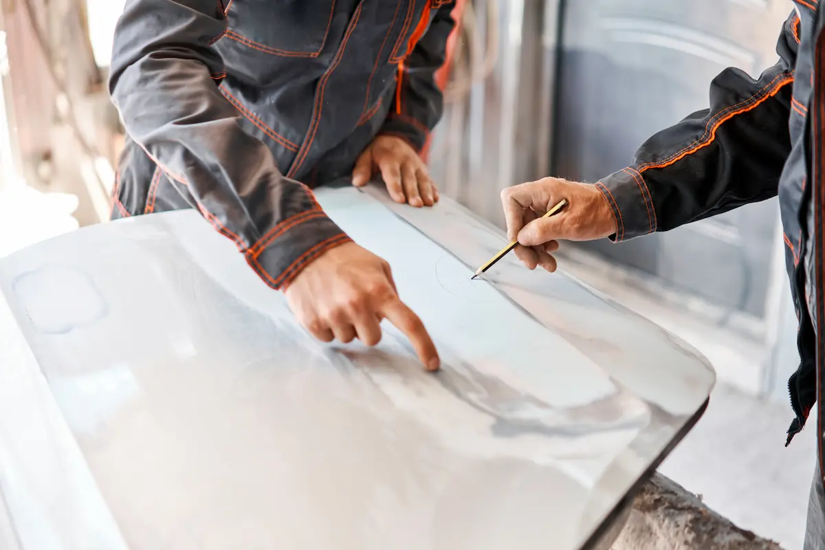 Two workers in dark gray jackets with orange stitching lean over a primed car panel, one pointing with a finger and the other marking the surface with a pencil.