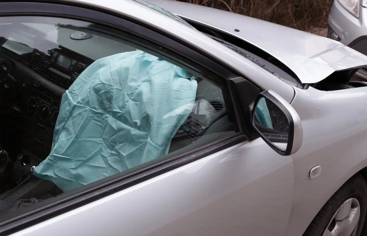 A silver car with a crumpled front fender and hood seen from the driver’s side, with a deployed light blue airbag filling the window behind a side mirror.