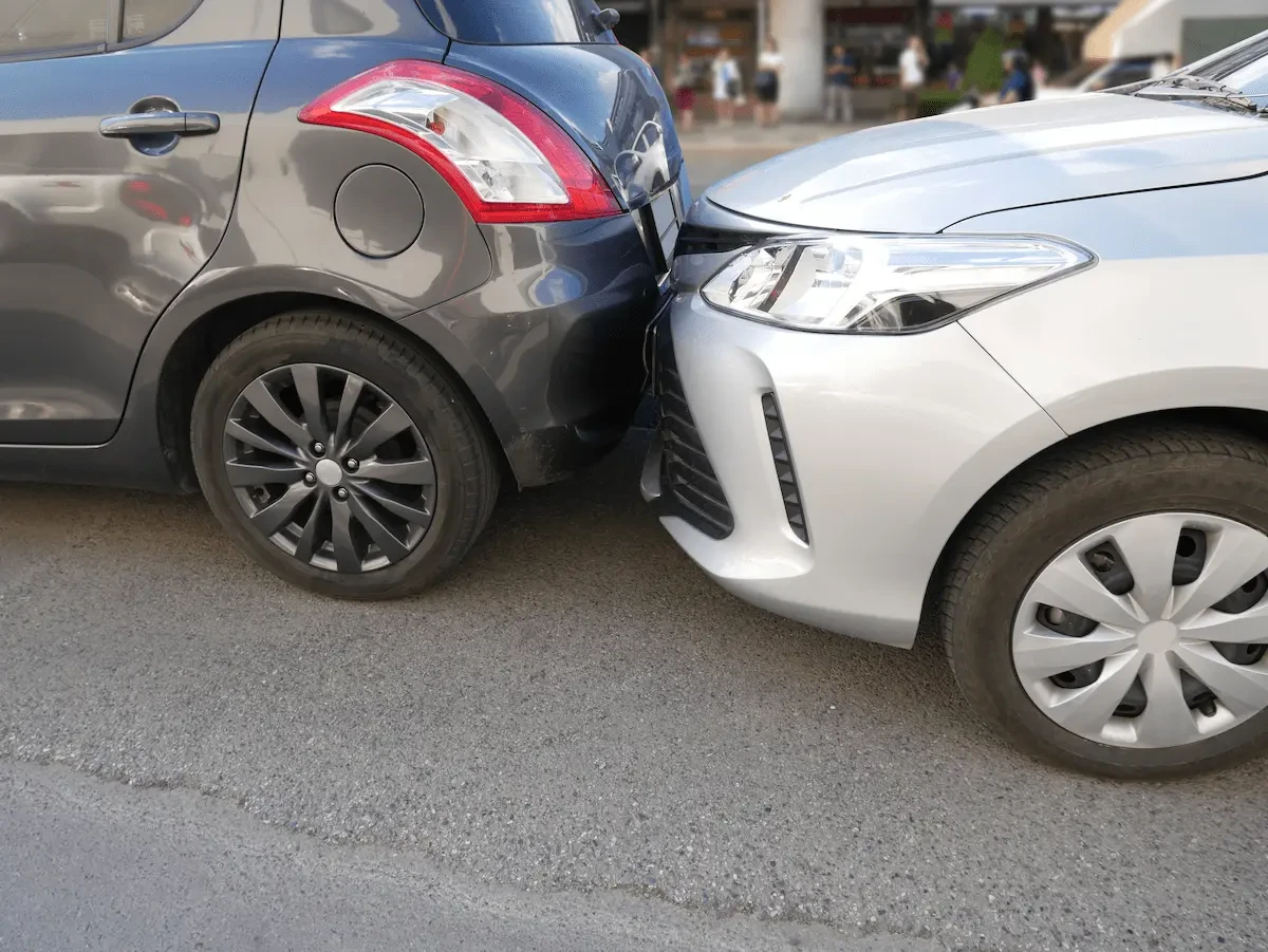 Alt text: The front of a silver sedan, its bumper cover pushed in and separated near the grille, presses against the rear bumper of a gray hatchback on an asphalt street, with blurred pedestrians in the background.