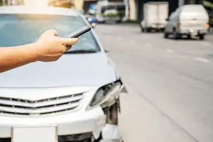 A hand holds a smartphone in front of a silver sedan with front-end damage on the driver’s side, parked at the edge of a city street.