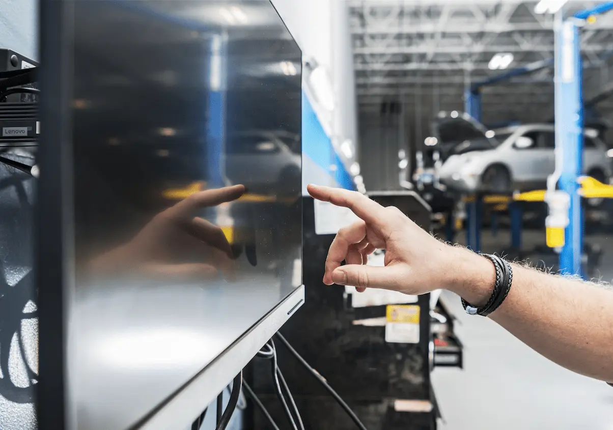 A person’s hand points at a computer monitor in an auto repair shop, with a silver car raised on a blue lift in the background.