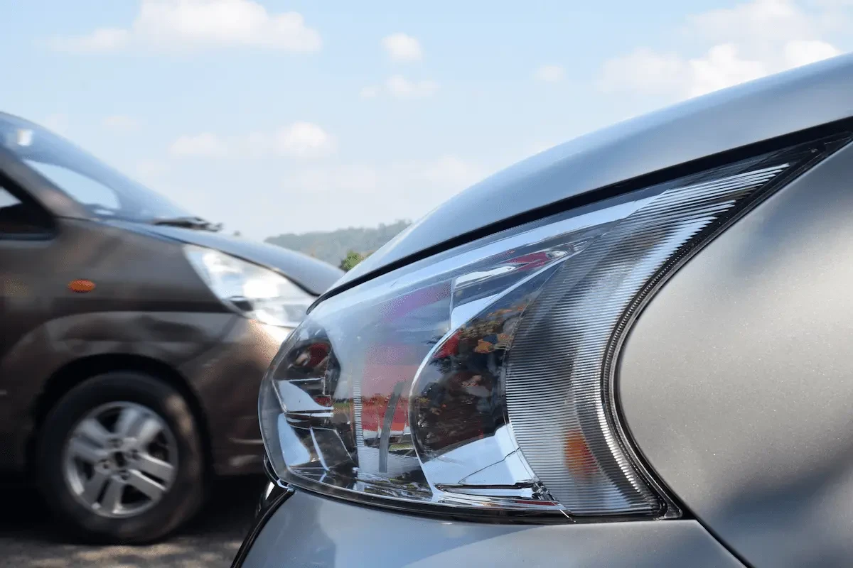 Close-up of the clear-lens headlamp assembly on a silver car, with an amber reflector strip along its inner edge, beside a blurred brown minivan parked under a partly cloudy sky with a tree-covered hill in the distance.