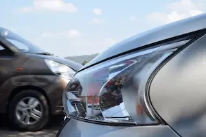 Close-up of the clear-lens headlamp assembly on a silver car, with an amber reflector strip along its inner edge, beside a blurred brown minivan parked under a partly cloudy sky with a tree-covered hill in the distance.