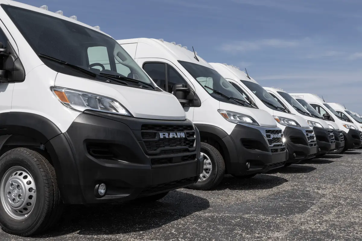 A diagonal row of white Ram ProMaster cargo vans on a gravel lot under a partly cloudy sky, with the nearest van’s black grille and “RAM” lettering in the foreground.