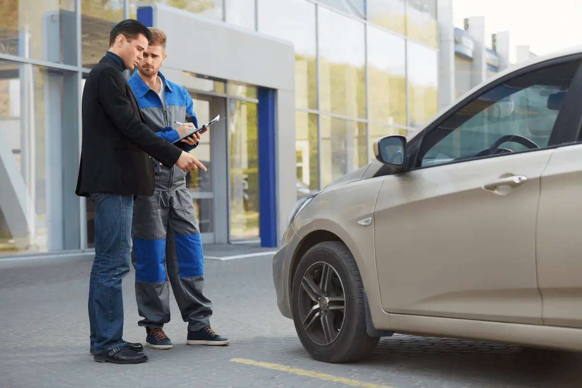 A man in a black blazer and jeans points toward the front of a tan sedan while a mechanic in blue and gray coveralls writes on a clipboard beside him, in front of a building with a glass facade.