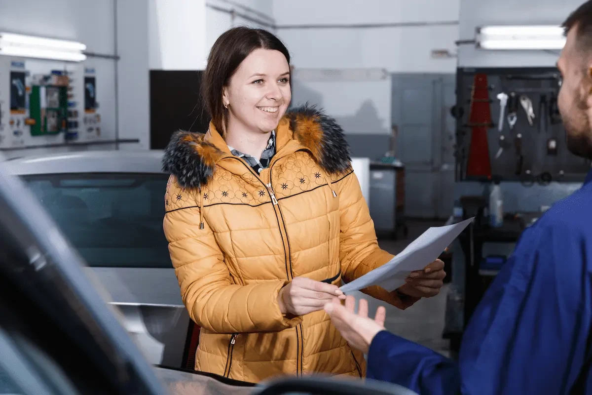 A smiling woman in an orange quilted winter jacket with a fur-trimmed hood hands a sheet of paper to a man in a blue work shirt inside a repair shop, with a silver car and tools on a pegboard wall in the background.