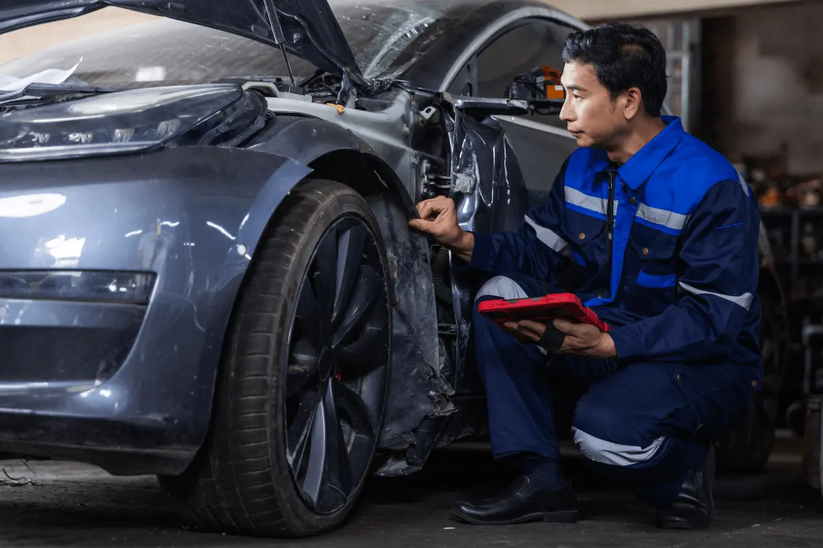 Technician kneeling beside a damaged gray sedan, holding a red tablet while inspecting the front wheel well area. The car’s hood is open and its windshield is cracked.