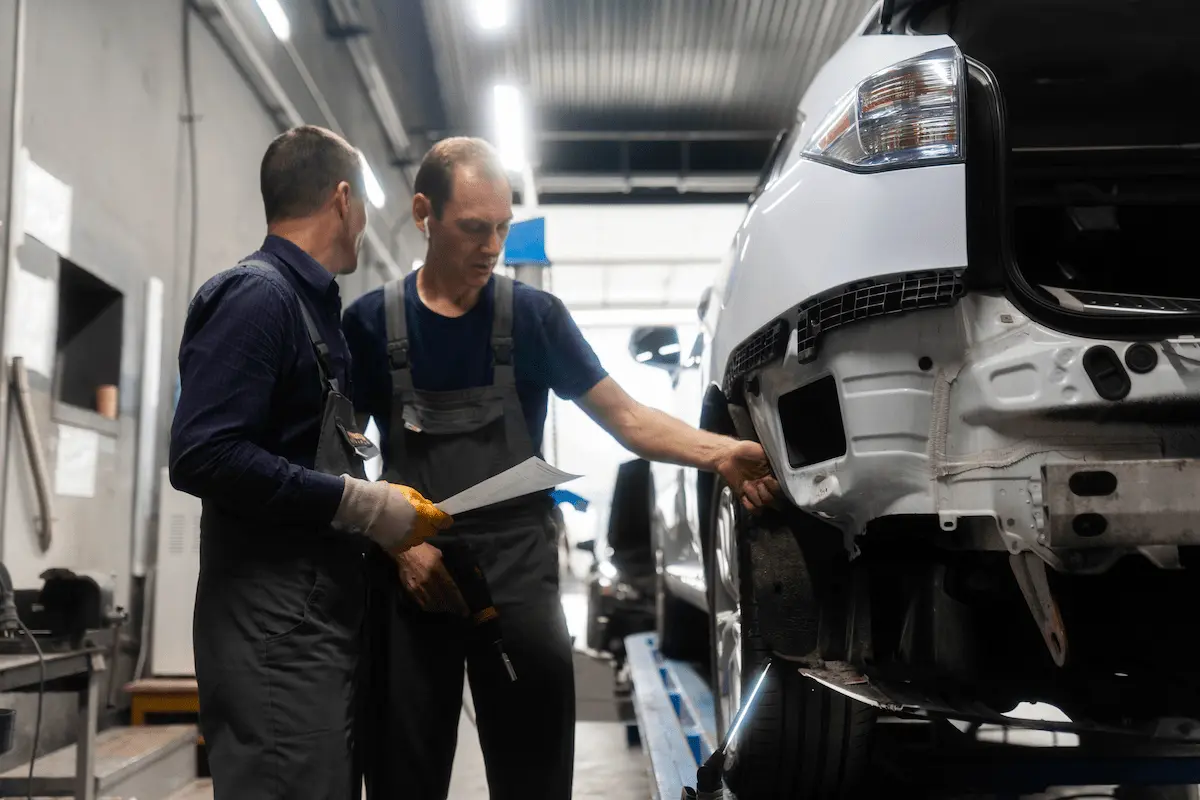 Two collision repair technicians in work overalls review a printed document beside a white vehicle on a lift, one pointing to damage where the rear bumper cover has been removed.
