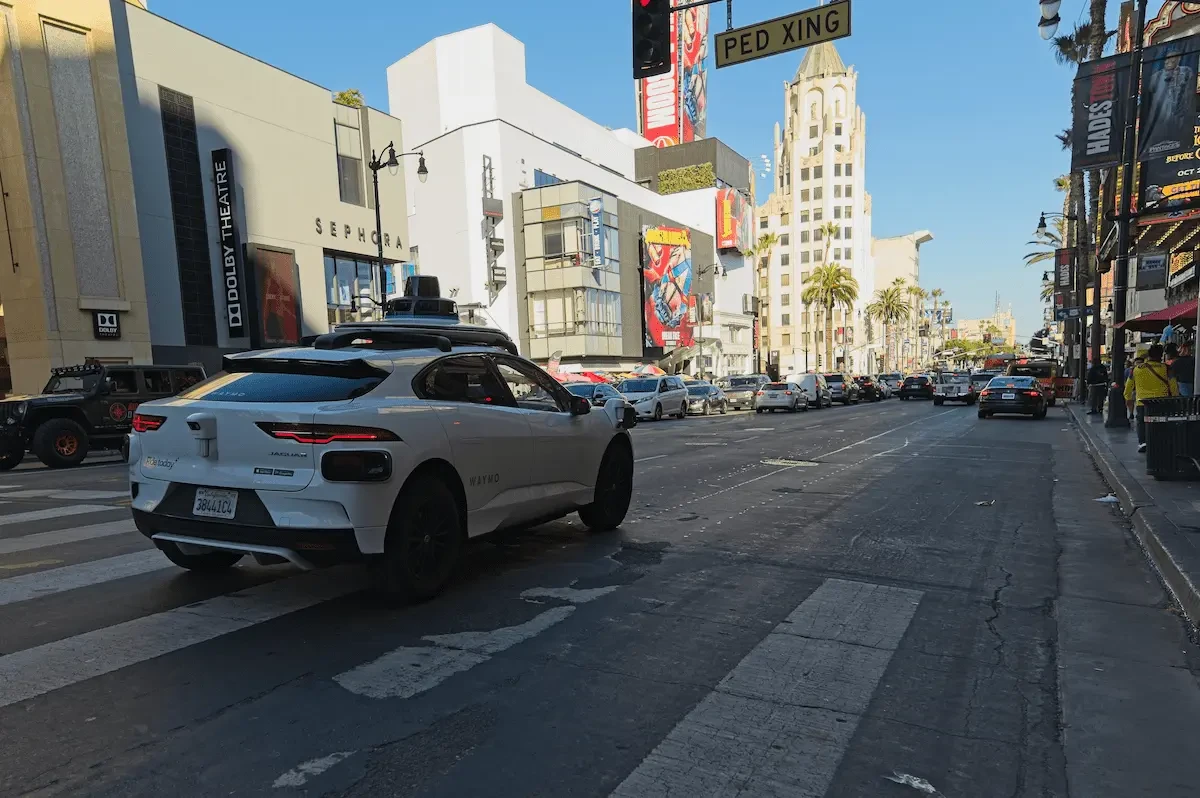 A white Jaguar SUV with Waymo lettering and a roof-mounted sensor unit sits at a crosswalk on a sunny city street, with a Dolby Theatre sign, a Sephora storefront, a "PED XING" sign, palm trees, and a line of traffic in the background.