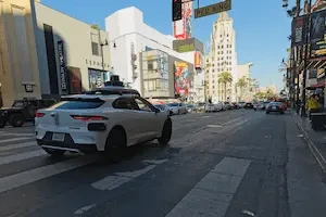 A white Jaguar SUV with Waymo lettering and a roof-mounted sensor unit sits at a crosswalk on a sunny city street, with a Dolby Theatre sign, a Sephora storefront, a "PED XING" sign, palm trees, and a line of traffic in the background.