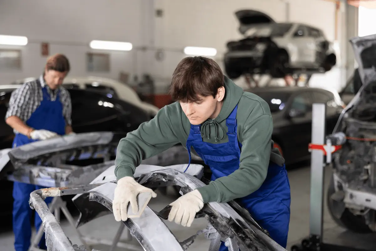 A young man in a green hoodie, blue bib overalls, and white work gloves leans over a gray bumper cover on a stand, holding a sanding block and a dark hand tool against it. Behind him, an older man in a plaid shirt and blue overalls works on another bumper. Vehicles fill the shop, including one raised on a lift.