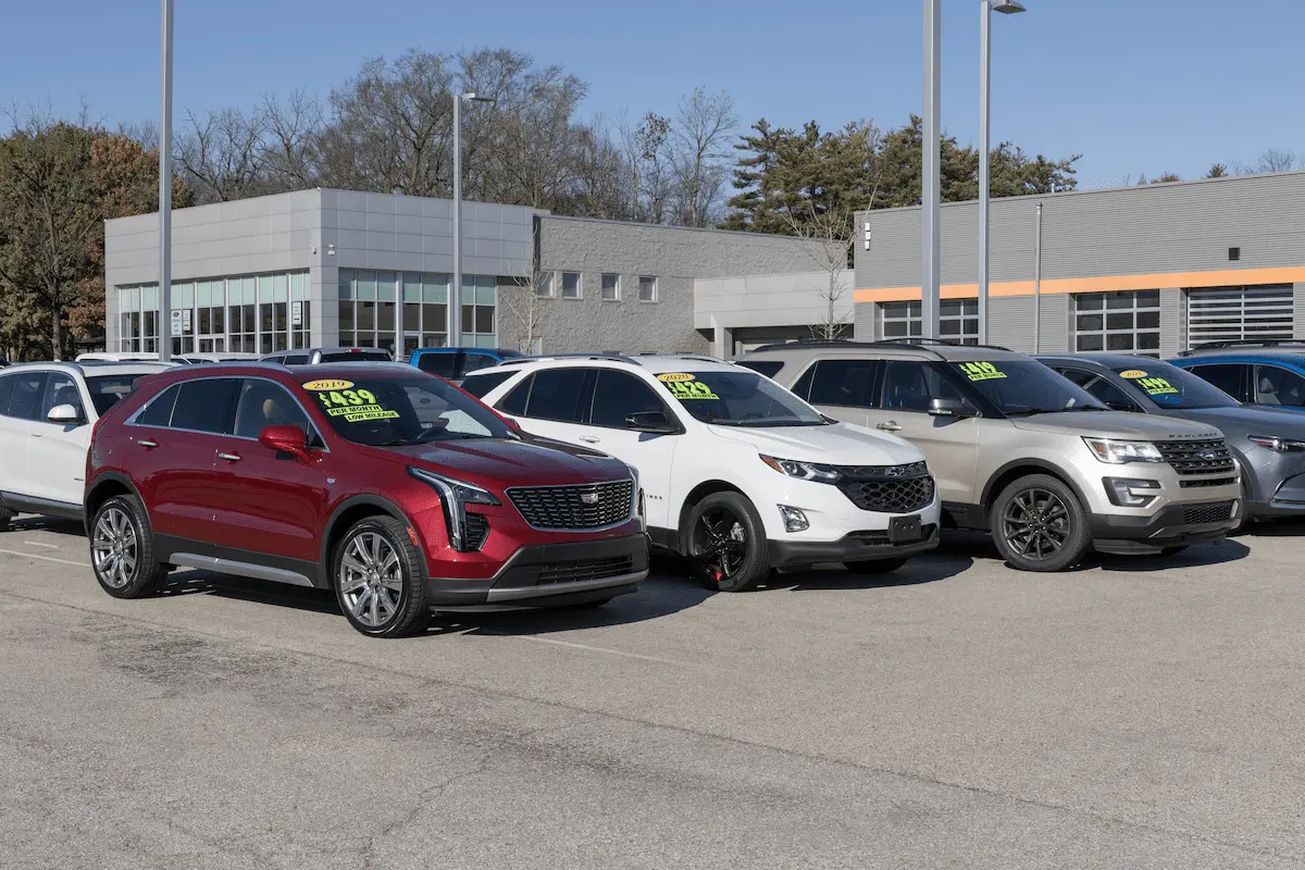 A red Cadillac SUV, a white Chevrolet SUV, and a silver Ford SUV parked in a row on a dealership lot, with yellow and green monthly price stickers on their windshields, in front of a gray building with large windows under a blue sky.