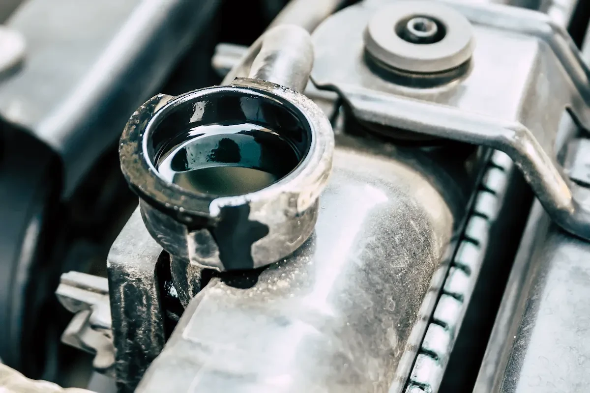 Close-up of an open radiator filler neck holding dark coolant, on a worn metal radiator with cooling fins and a mounting bracket inside an engine compartment.
