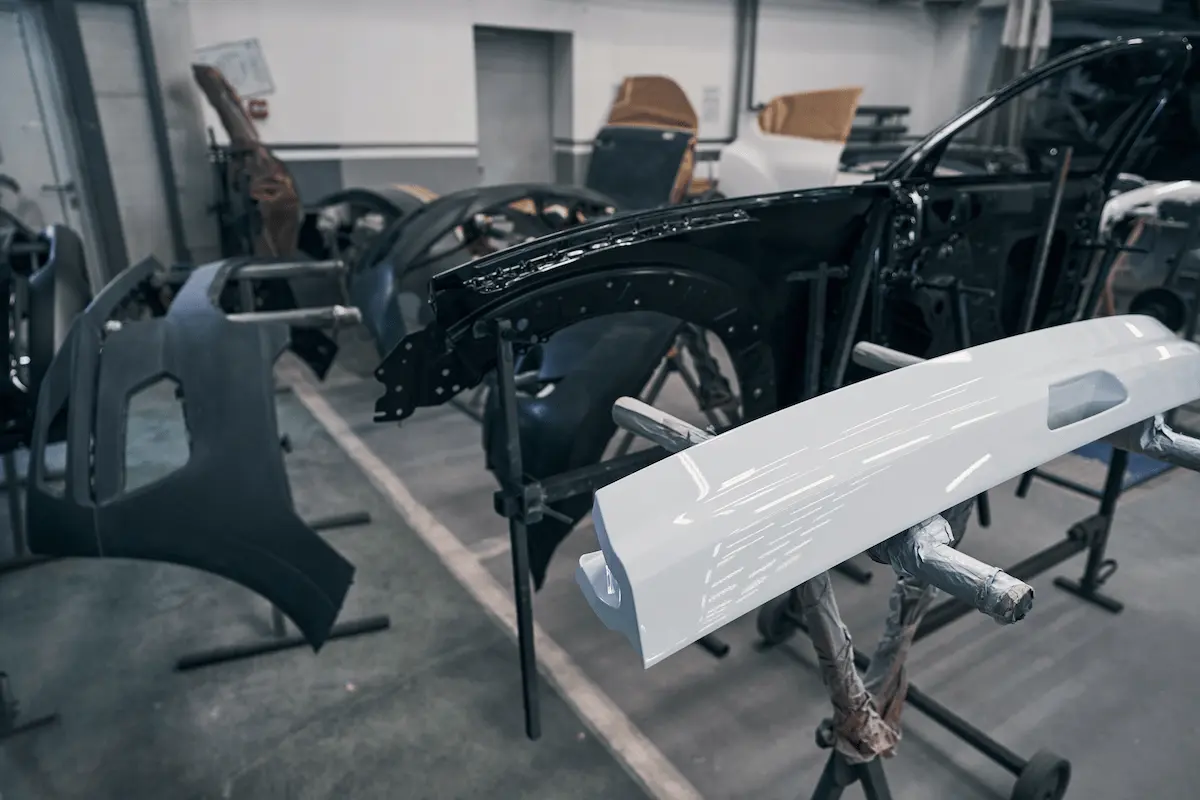 A black front fender, a glossy white bumper cover and a dark gray bumper cover mounted on metal stands in an auto body shop, with paper-masked panels and white walls in the background.